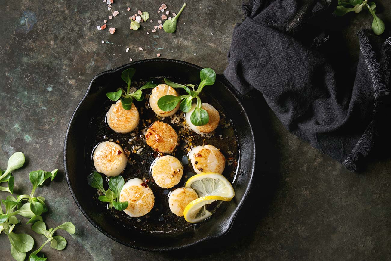 910509922 Fried scallops with butter lemon spicy sauce in cast-iron pan served with green salad and textile napkin over old dark metal background. Top view, space