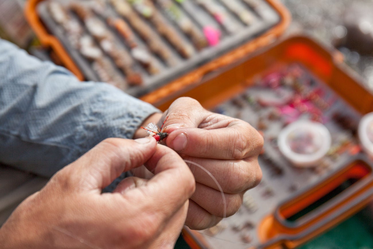 A close-up image of a man tying a fish fly. Tackle box in background. Fly-fishing theme.