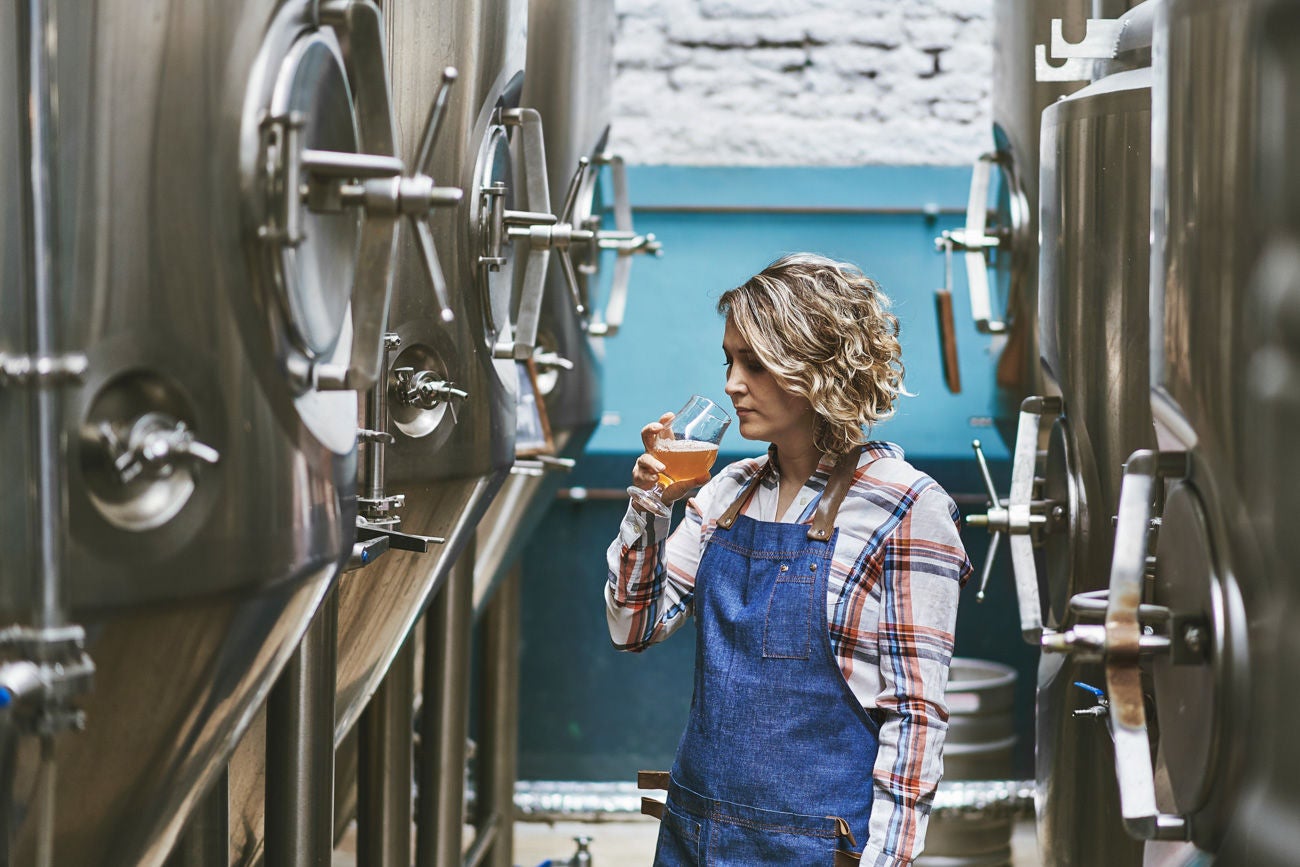 1197894094 Female Buenos Aires craft brewer in mid 30s standing between vats and examining beer sample in glass.