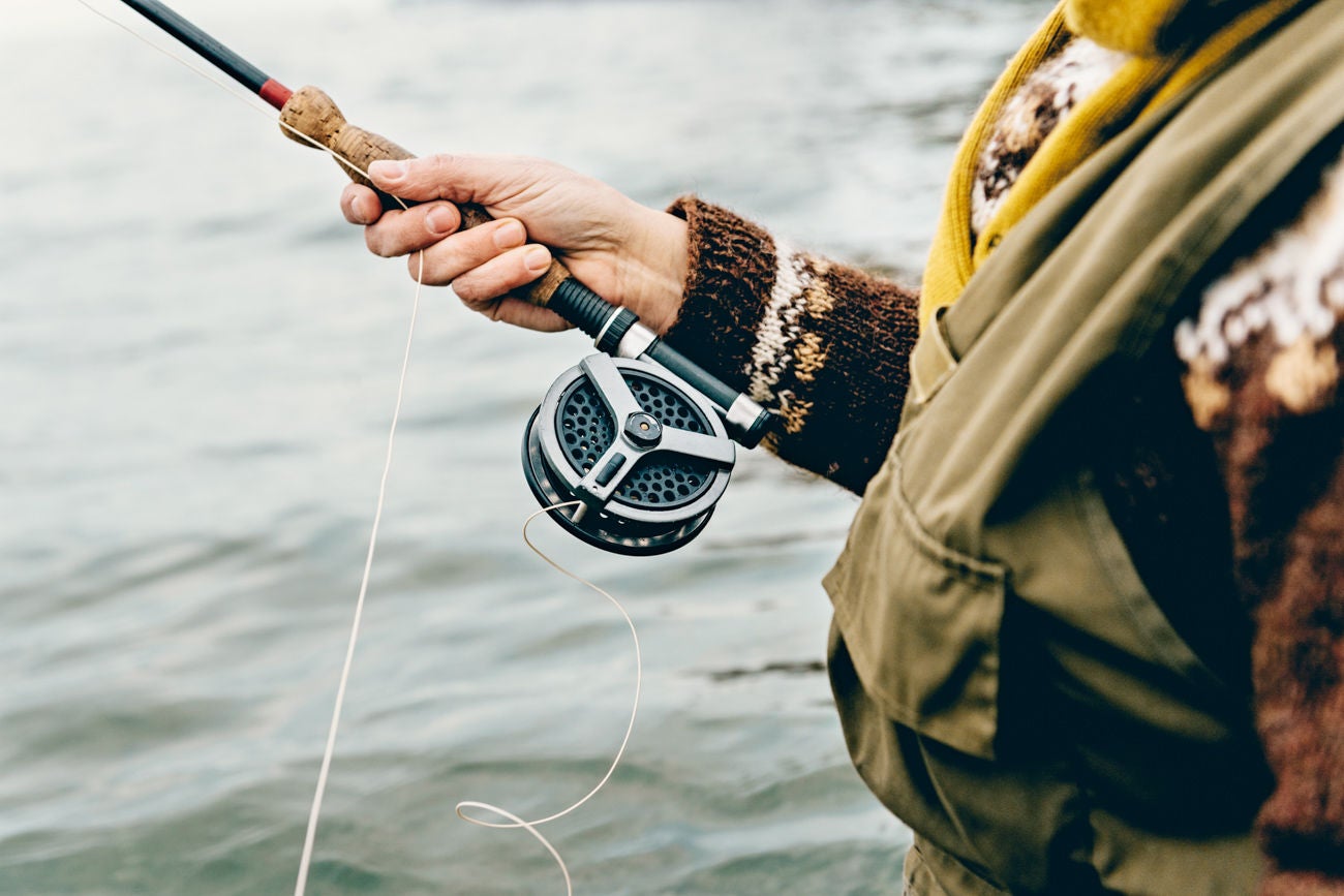 Close up portrait of a  fly fisherman's hands as she works the line and reel. She is wearing a green cotton gillet,waders, a patterned sweater and a mustard coloured scarf. Colour, horizontal format photographed against a calm sea with lots of copy space. Photographed on location at Nordfeld Strand on the island of MÃ¸n in Denmark.