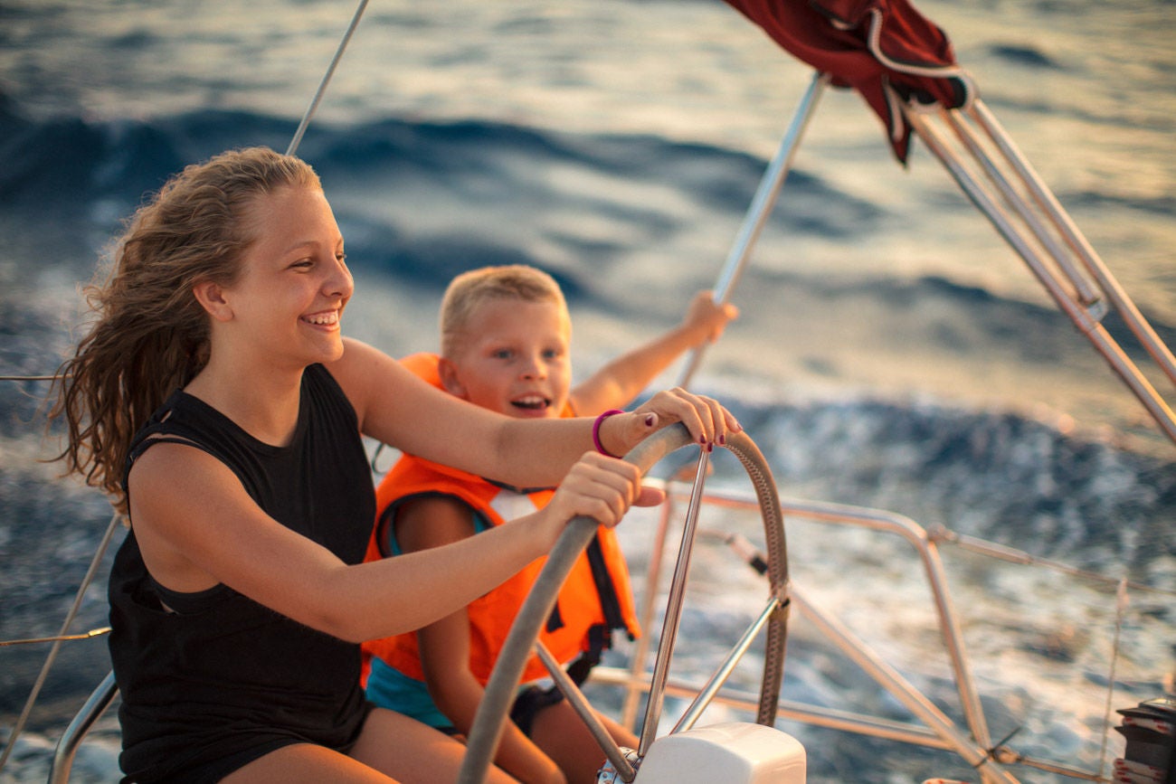 Teenage girl sitting with her younger brother, enjoying at summer and having a fun while steering with yacht at sunset. They are very excited and happy